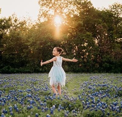 Girl in a blue dress with arms outstretched in a field of blue wildflowers, sun shining through trees.