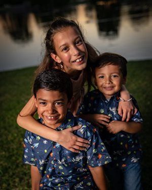 Three smiling children pose outdoors by a pond; one girl hugs two boys, all wearing patterned blue shirts.