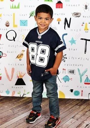 Young child in a Dallas Cowboys jersey and jeans, smiling, standing in front of a patterned backdrop.