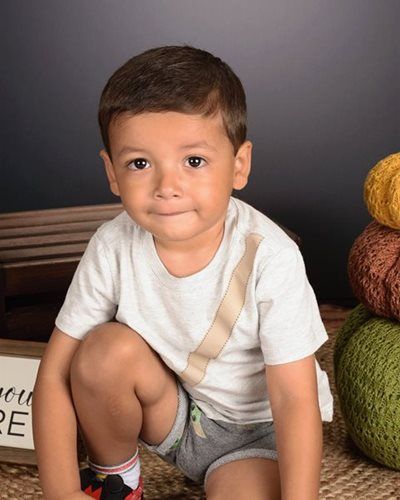 Boy in light shirt and shorts kneels, smiling, near pumpkins.