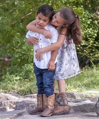 Girl hugging boy, both wearing cowboy boots, near water. Boy looks uncertain, girl smiles. Outdoors.