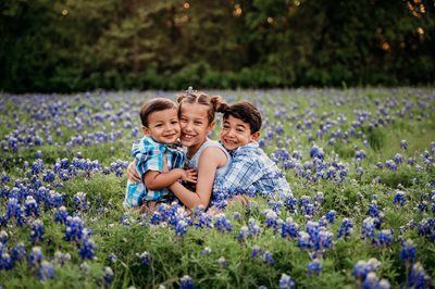 Three smiling children embrace in a field of blue wildflowers.