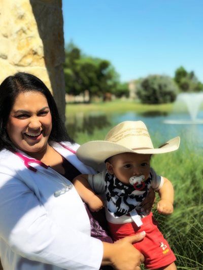 Woman in white coat holds baby in cowboy hat; fountain and pond in background.