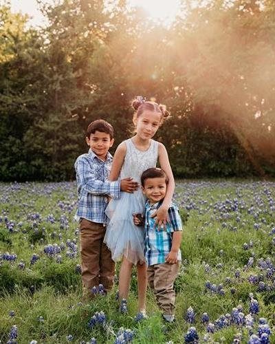 Three children in a field of blue flowers. Girl hugs two boys; sun shines through trees.