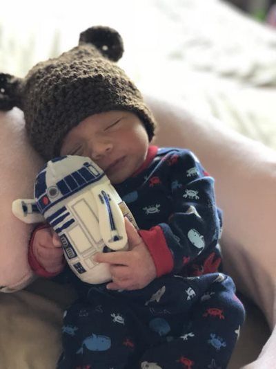Newborn wearing a bear hat and Star Wars pajamas, asleep while cuddling a R2-D2 toy.