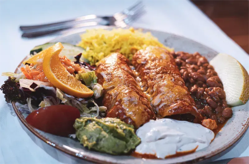 Ein Teller mit Enchiladas, Reis, Bohnen, Salat, Guacamole, Sauerrahm und einer Melonenscheibe.