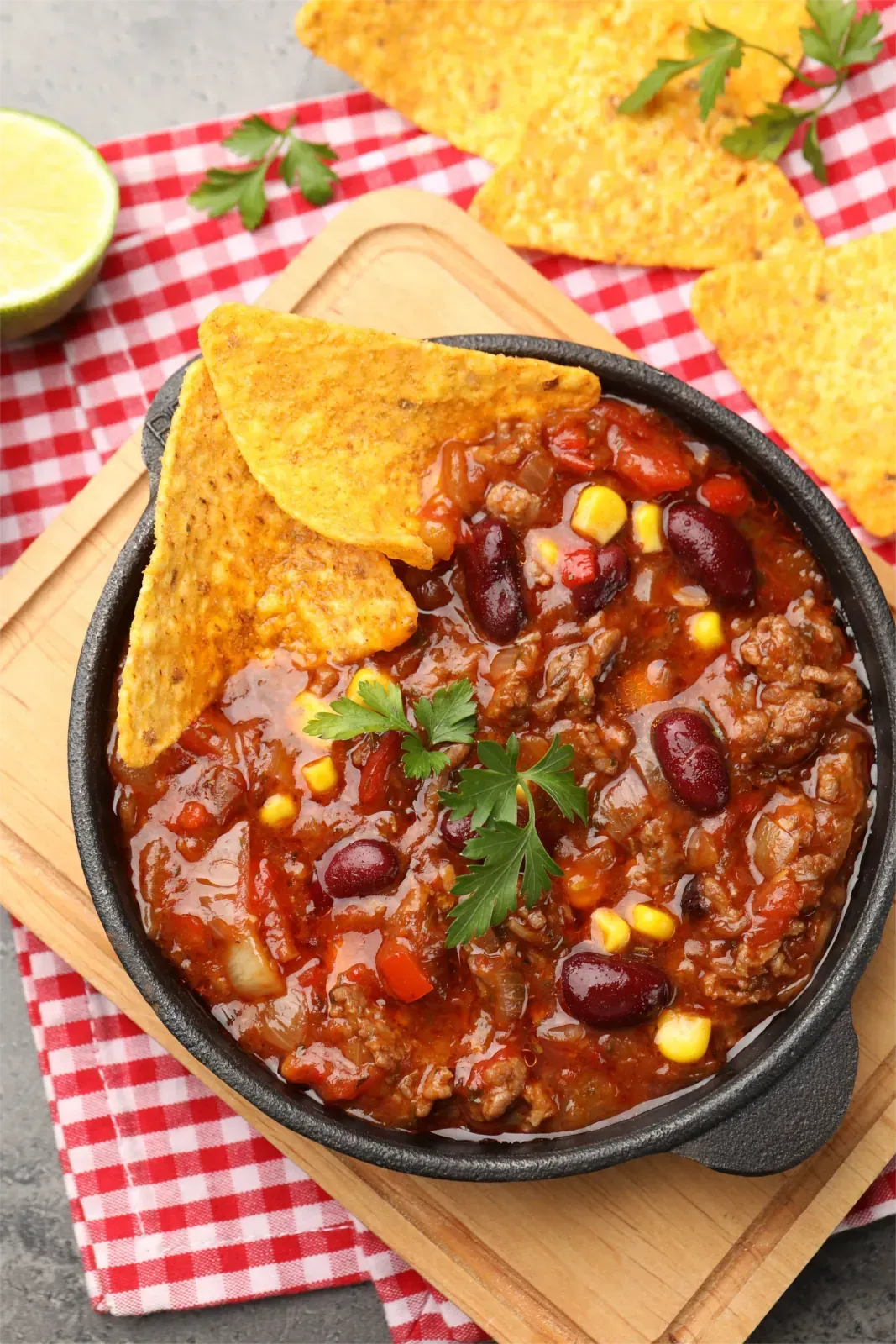 Chili mit Bohnen und Mais in einer schwarzen Schüssel, Tortilla-Chips, Petersiliengarnitur, Limette auf einem rot-karierten Tuch.
