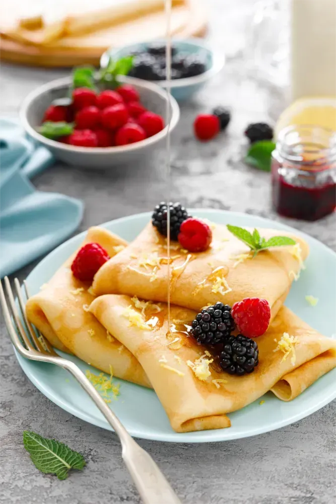 Crêpes mit Beeren, Zitronenschale und Honig auf einem blauen Teller, im Hintergrund weitere Beeren und Toppings.