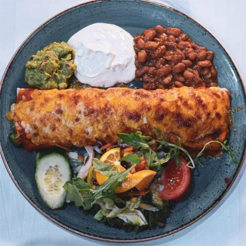 Teller mit mexikanischem Essen: Enchilada, Bohnen, Guacamole, Sauerrahm, Salat und eine Gurkenscheibe.