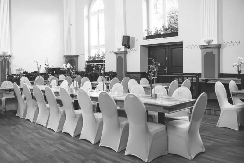 Festsaal mit langen, für ein Essen gedeckten Tischen. Weiße Stuhlhussen, hohe Fenster und Pflanzen.