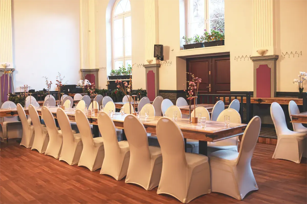 Lange, rechteckige Tische stehen in einem großen Raum mit weißen Stühlen. Holzboden und hohe Fenster.