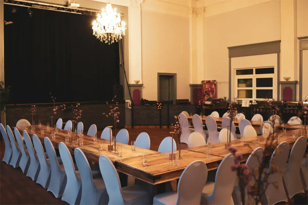 Großer Festsaal mit U-förmig gedeckten Tischen für ein Abendessen, Bühne im Hintergrund.