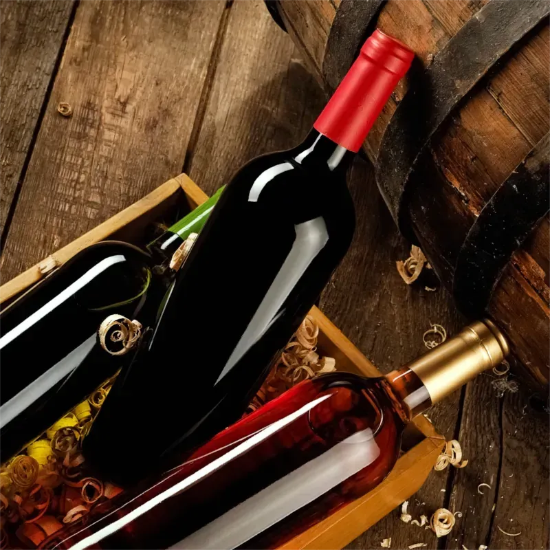 Weinflaschen in einer Holzkiste neben einem Fass. Dunkelrote, goldene und grüne Flaschen auf Holz.