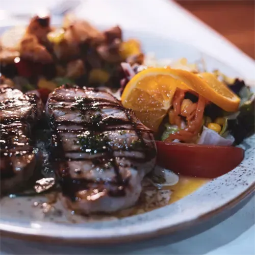 Gegrilltes Fleisch mit Salat, Tomaten und Orangenscheiben auf einem weißen Teller.