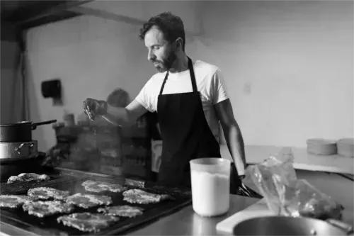 Ein Koch in schwarzer Schürze würzt Speisen auf einer Grillplatte in einer Küche.