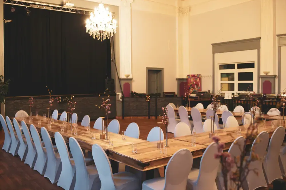 Der Festsaal ist für die Veranstaltung hergerichtet, Tische mit weißen Stühlen sind um die Bühne herum angeordnet.