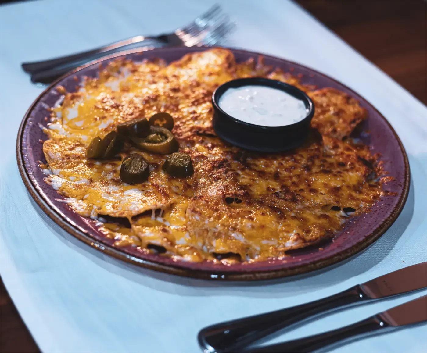 Nachos mit geschmolzenem Käse, Jalapeños und einer Beilage weißer Dip-Sauce auf einem violetten Teller.