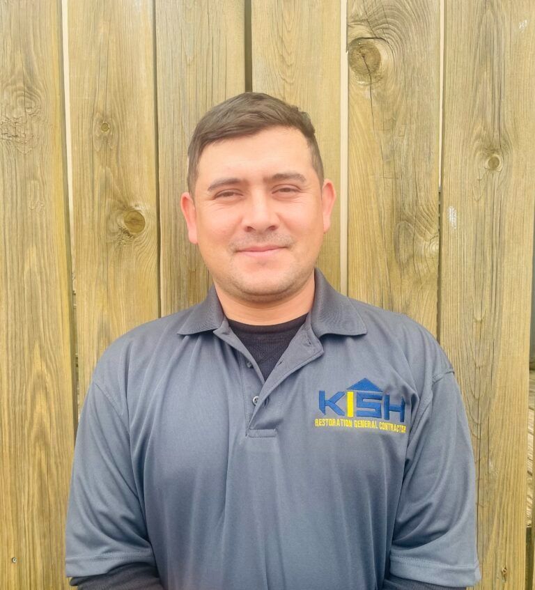 A man wearing a grey shirt with the word kash on it is standing in front of a wooden fence.