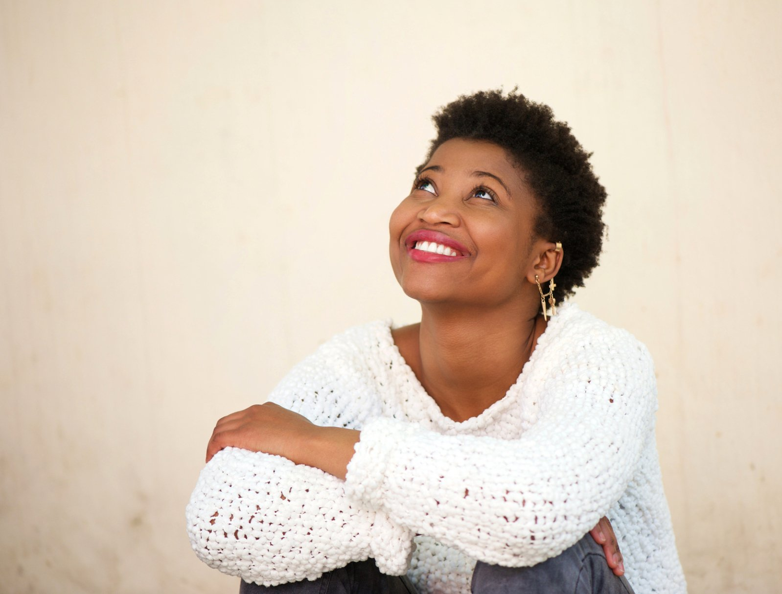 A woman in a white sweater is sitting on the floor with her arms crossed and smiling.