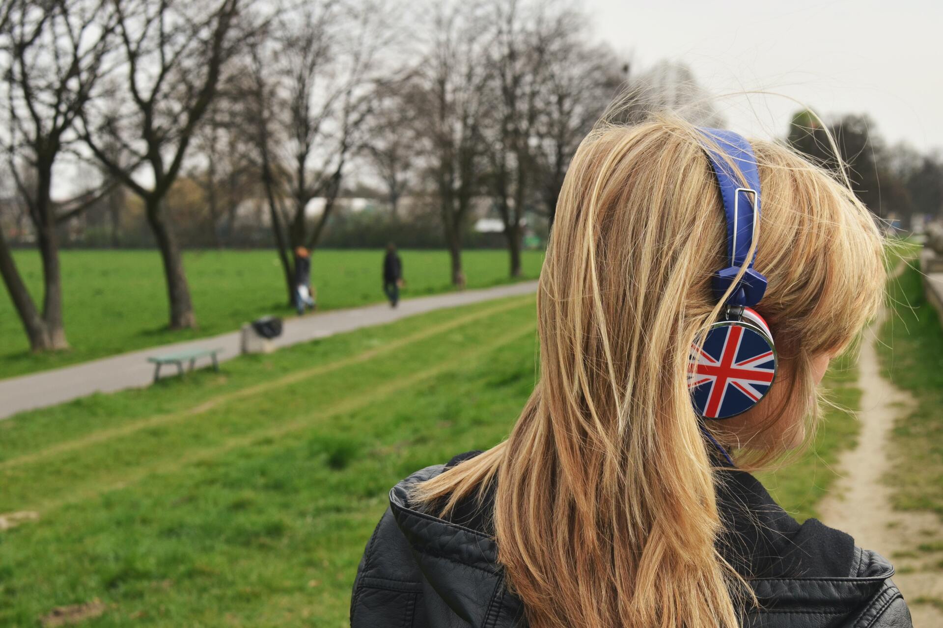 A woman wearing headphones with the british flag on them is standing in a park.