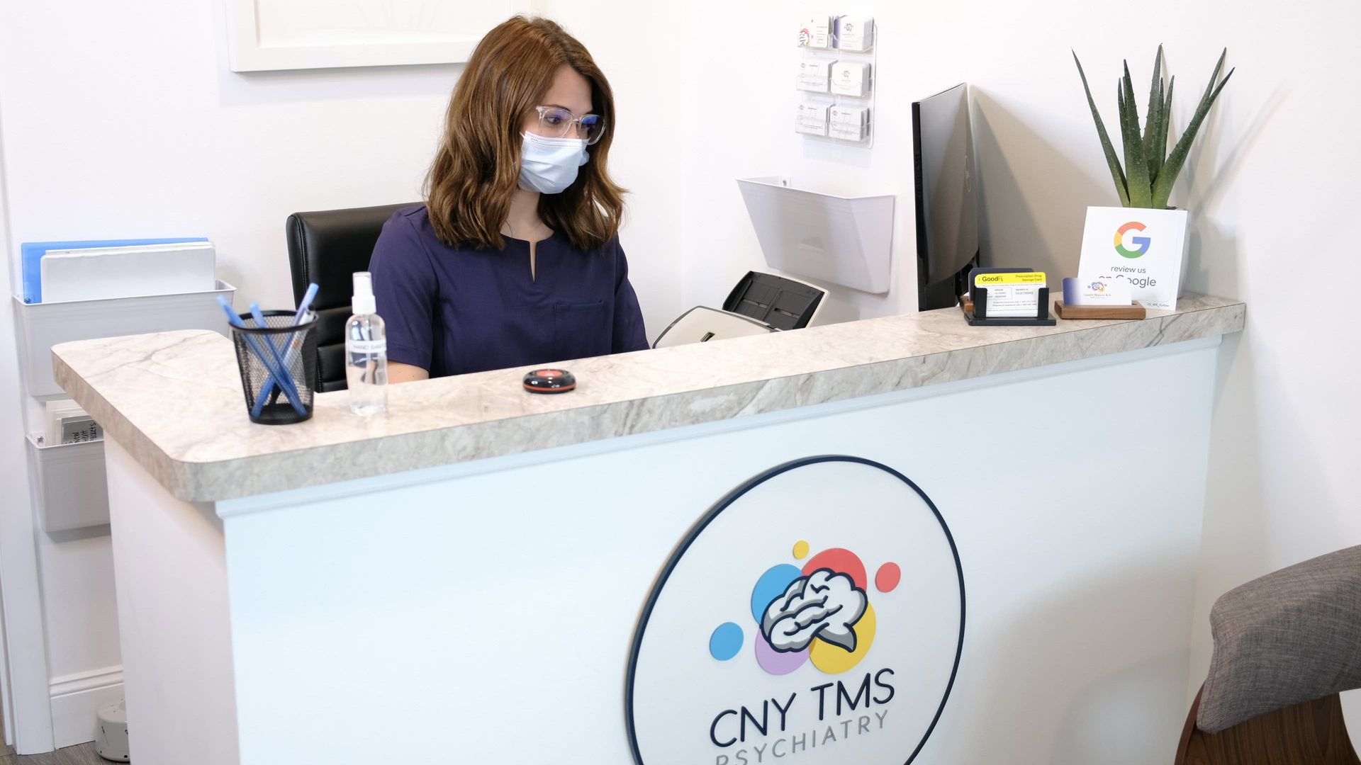A woman wearing a mask is sitting at a reception desk.