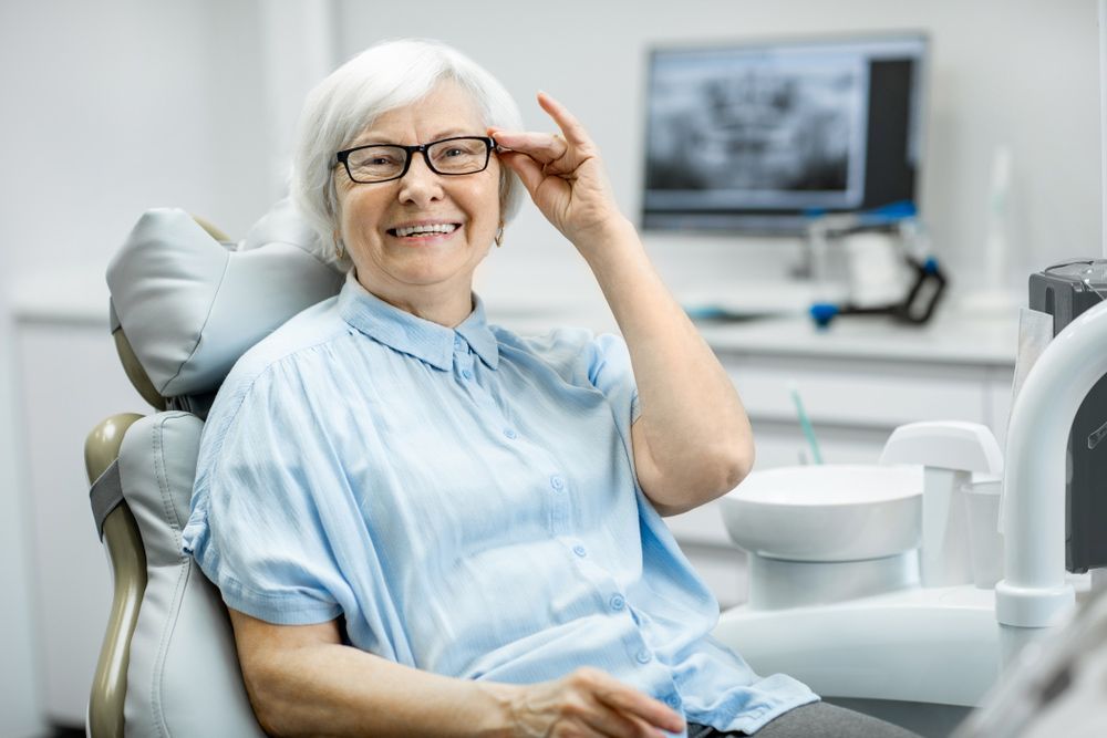 An Elderly Woman is Sitting in a Dental Chair Adjusting Her Glasses — Complete Denture Clinic in Kanwal, NSW