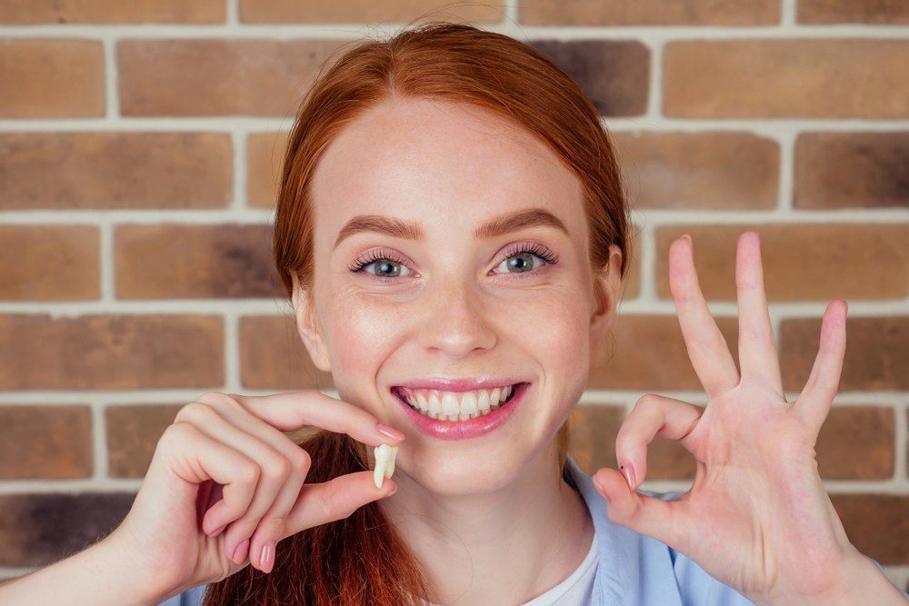 A Woman is Holding a Tooth in Her Hand and Giving an Ok Sign — Complete Denture Clinic in Kanwal, NSW