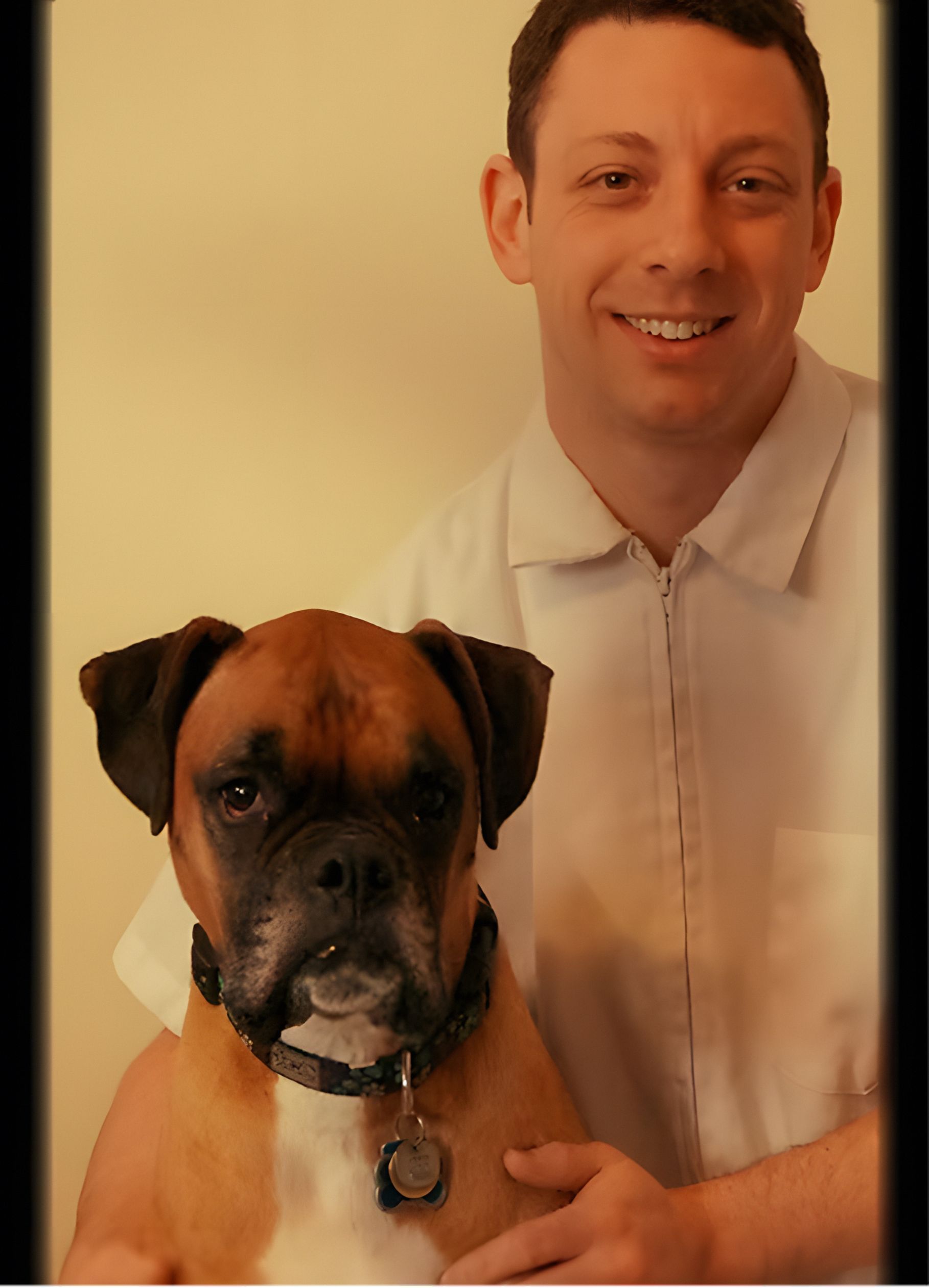 Man smiling with a boxer dog. The dog sits in front, wearing a collar. Both are indoors.