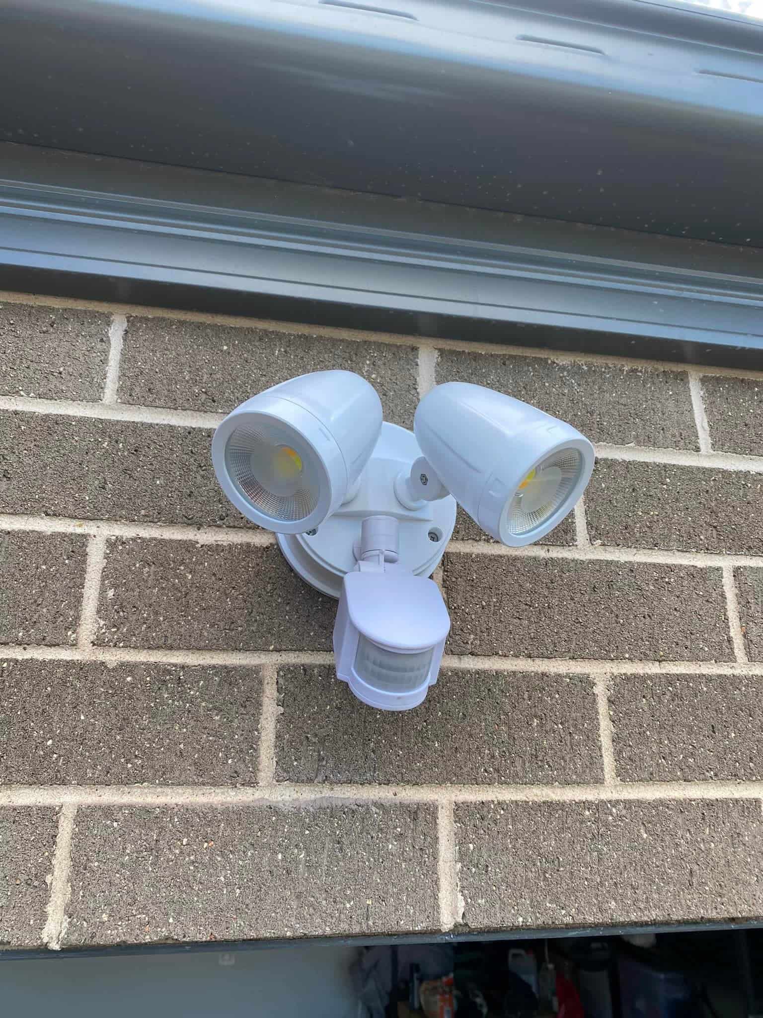 White Motion-sensing Floodlight on a Brick Wall Below a Gray Garage Door — SparkSafe Electrical in Dapto, NSW
