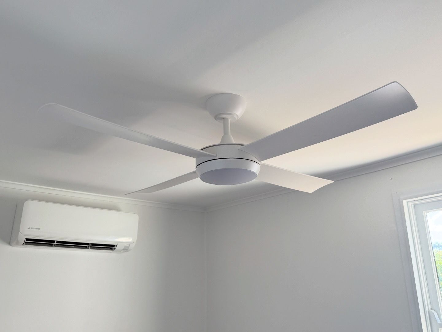 White ceiling fan with four blades, installed in a white room. An air conditioner is mounted on the wall  — SparkSafe Electrical in Dapto, NSW