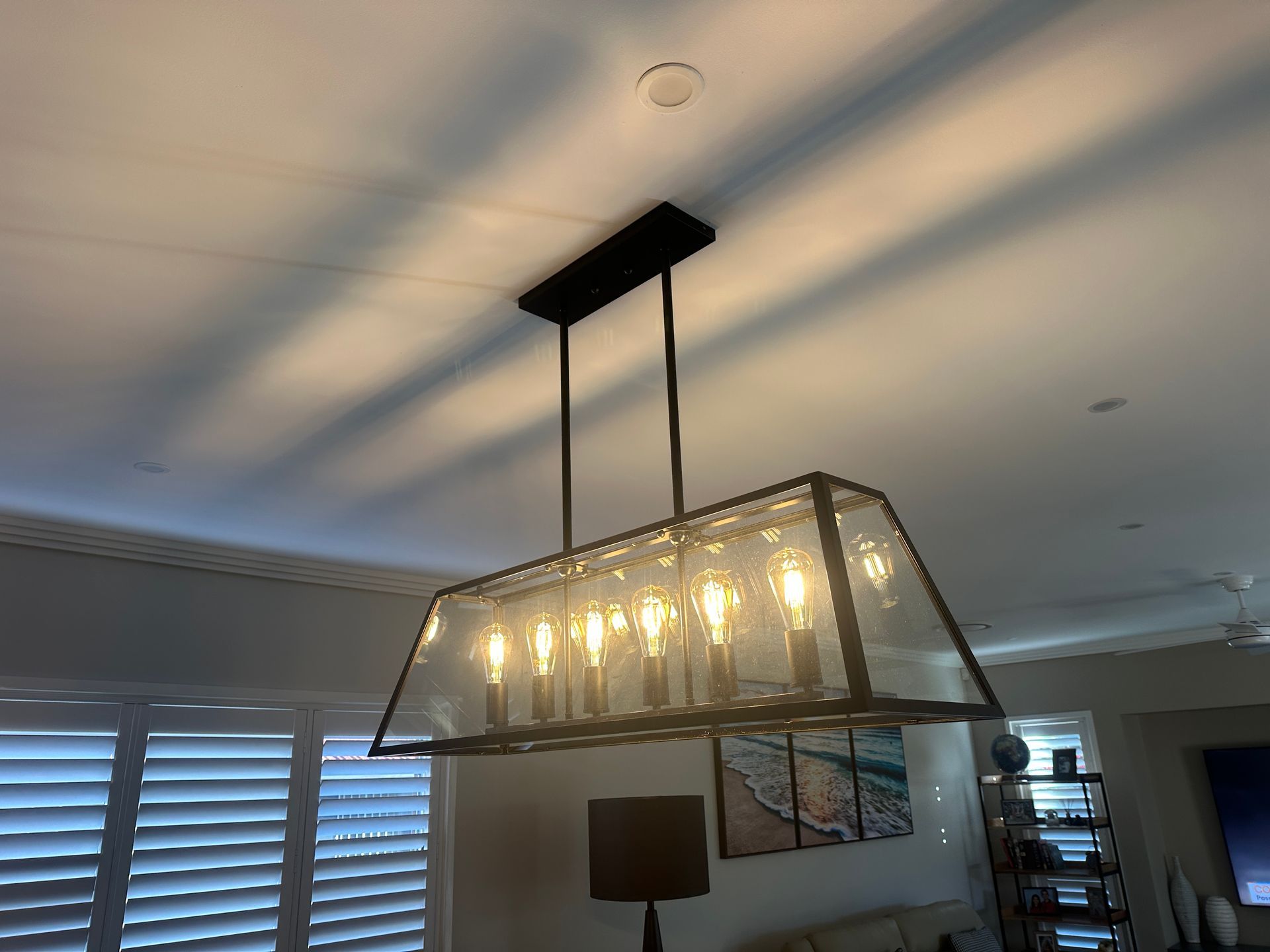 Rectangular chandelier with clear glass panels hangs from a black ceiling mount, illuminating a room with white shutters — SparkSafe Electrical in Gerringong, NSW