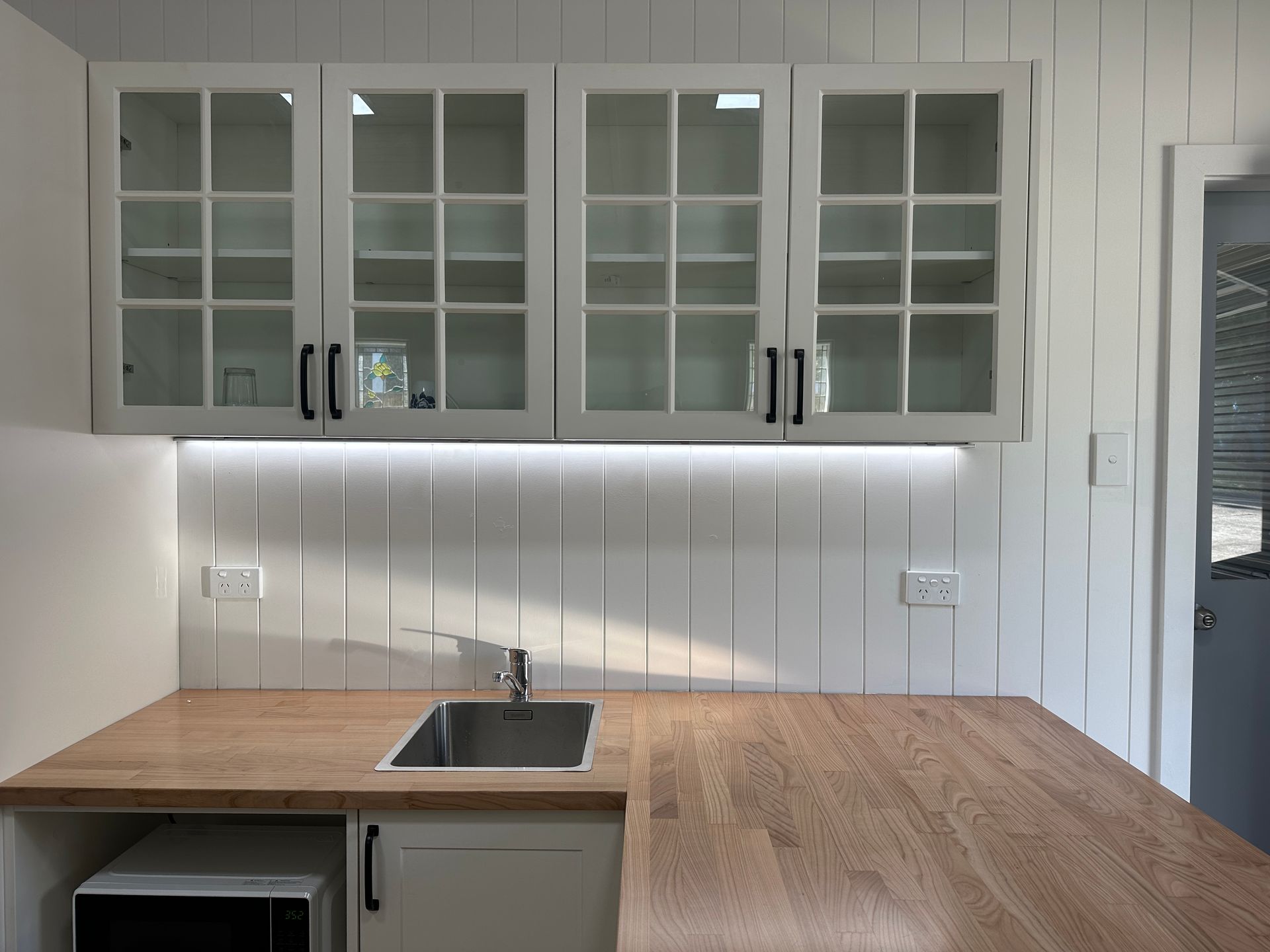 White kitchen cabinets with glass doors over a wood countertop, sink, and backsplash, with under-cabinet lighting  — SparkSafe Electrical in Dapto, NSW