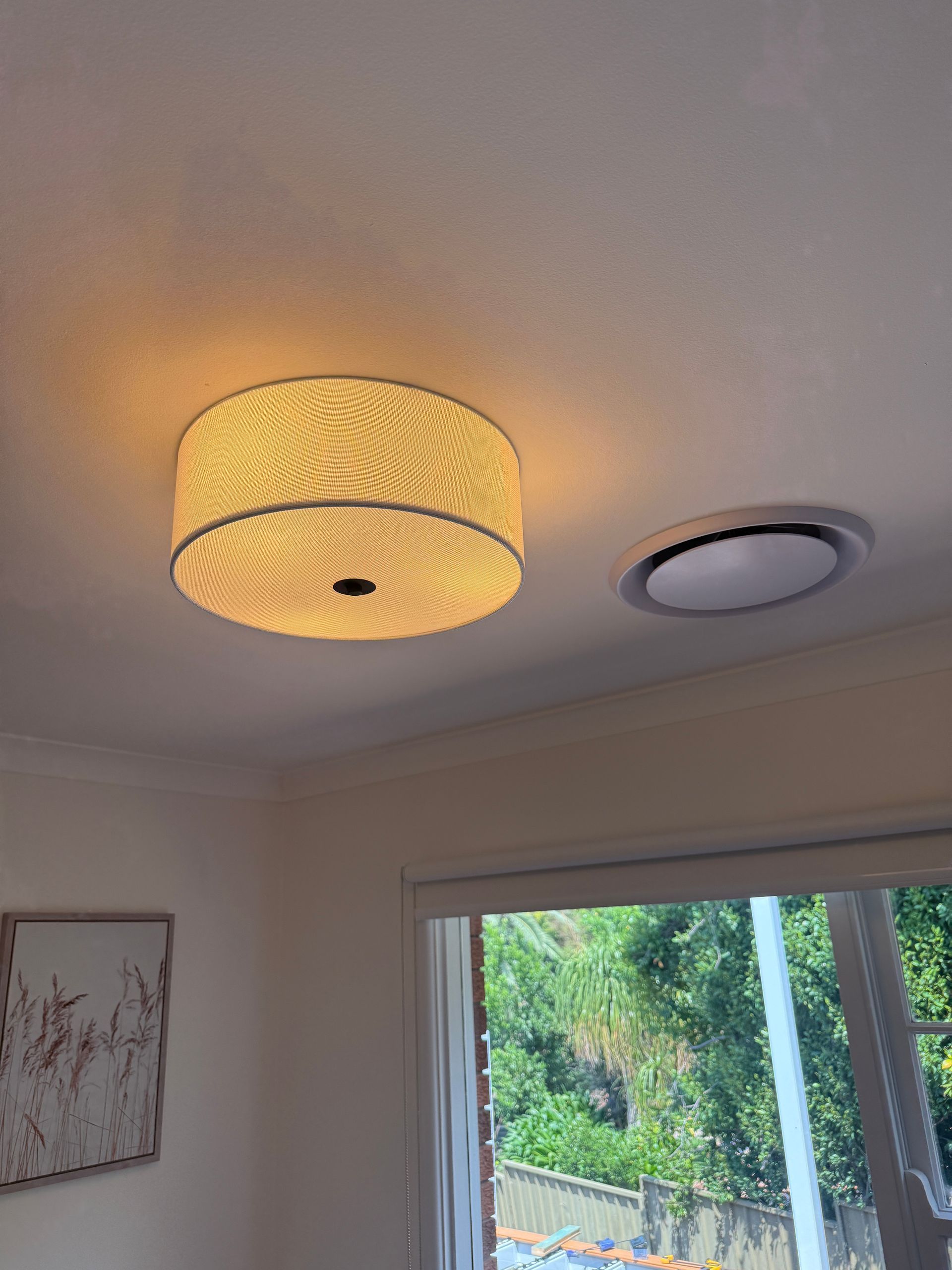 A round, illuminated ceiling light fixture next to a smaller, recessed ceiling vent, near a window  — SparkSafe Electrical in Dapto, NSW