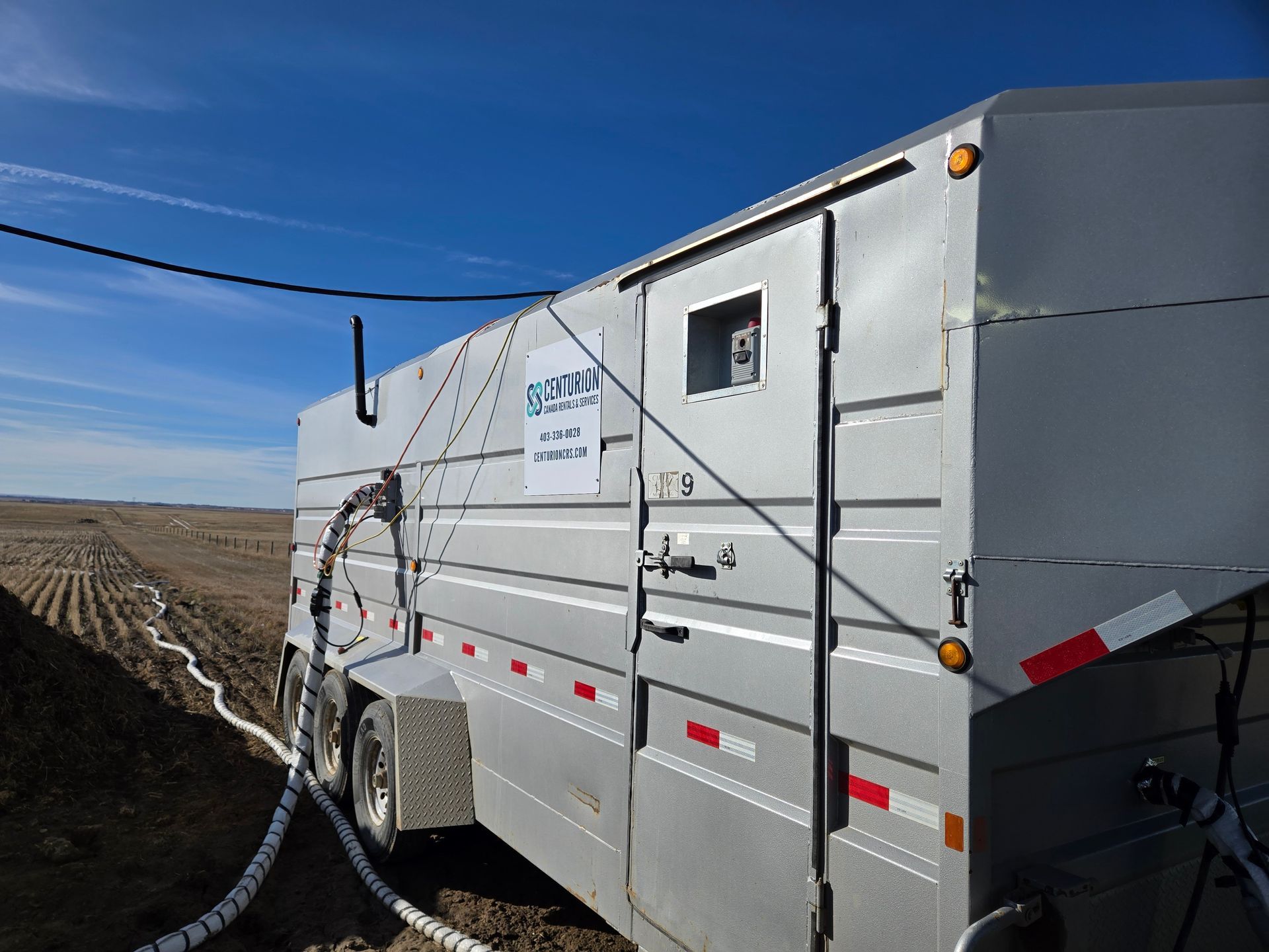 Silver trailer in a field, attached to white hoses. Blue sky in the background.