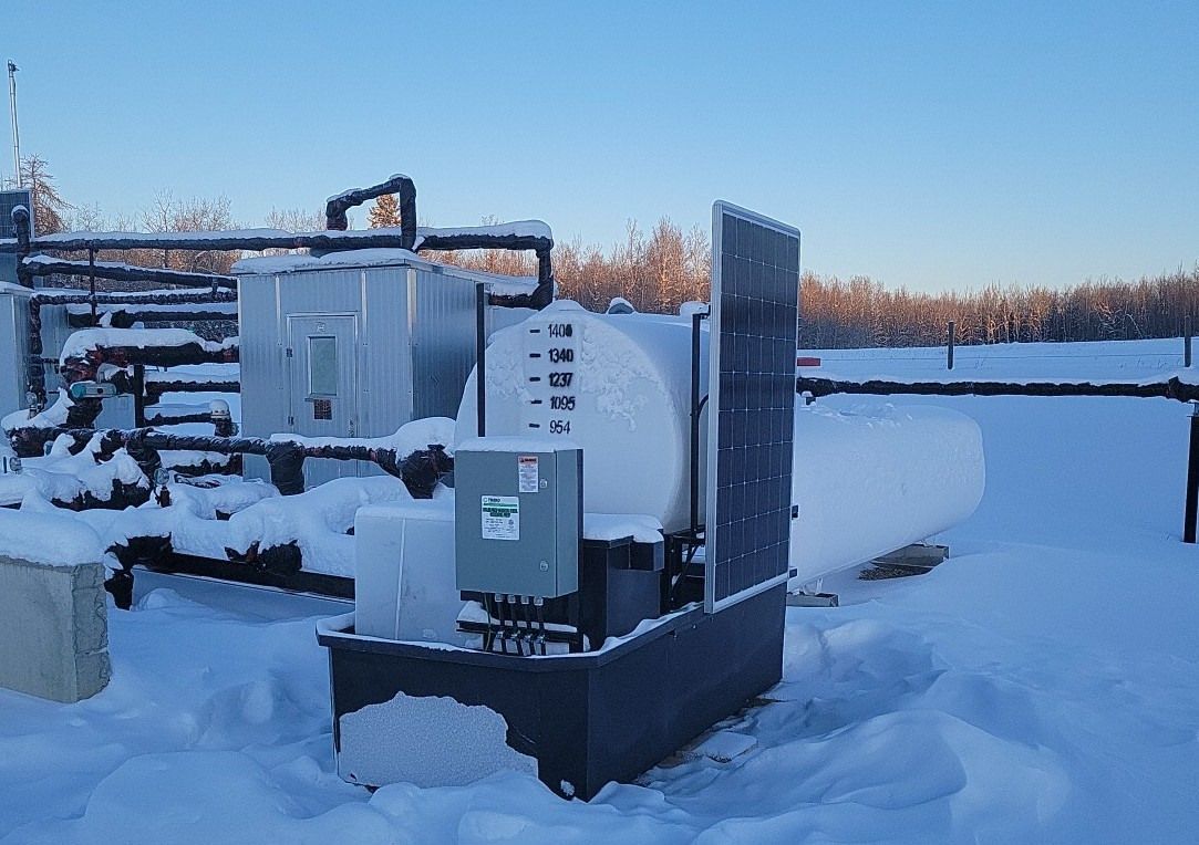 Snow-covered industrial equipment outdoors: a tank, control box, and solar panels against a winter landscape.