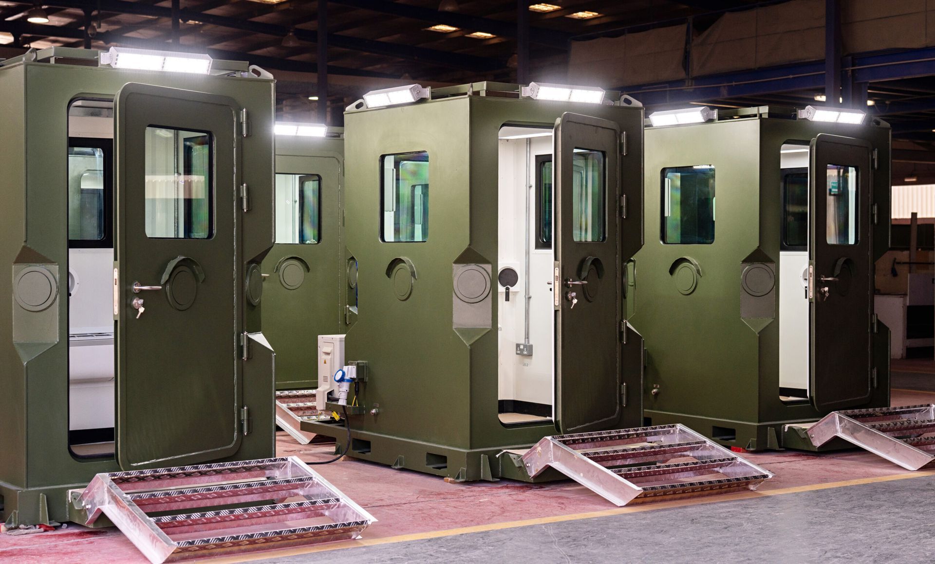 Three olive-green, boxy structures with open doors and ramps, possibly for accessibility, under bright lights.