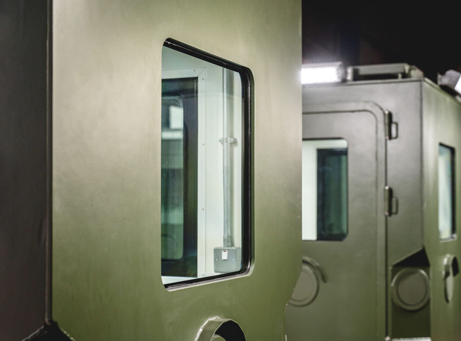 Olive-green armored vehicle exterior with rectangular window, door, and industrial setting.