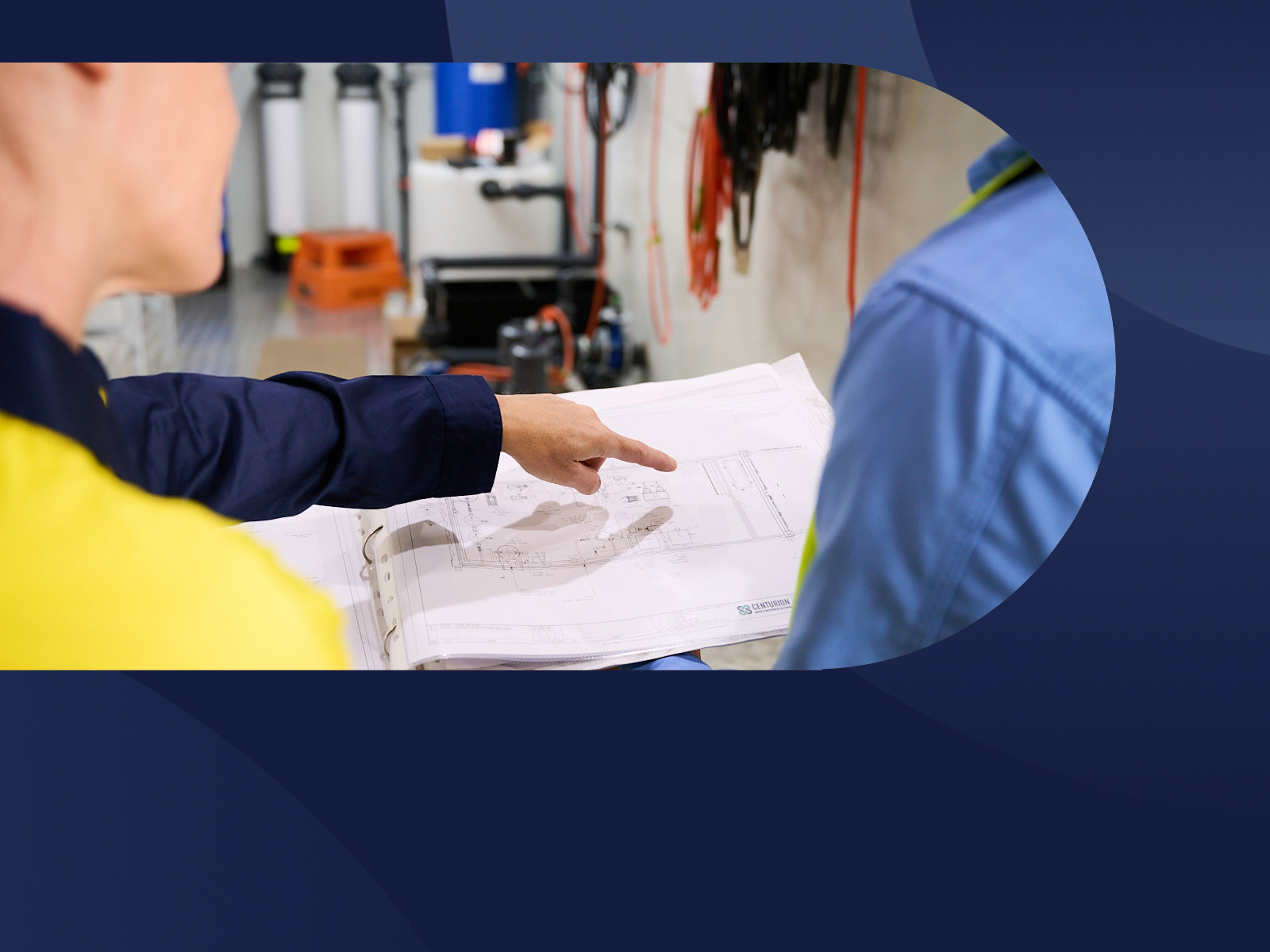 Two people reviewing plans on a workbench in a workshop, one pointing at a blueprint.