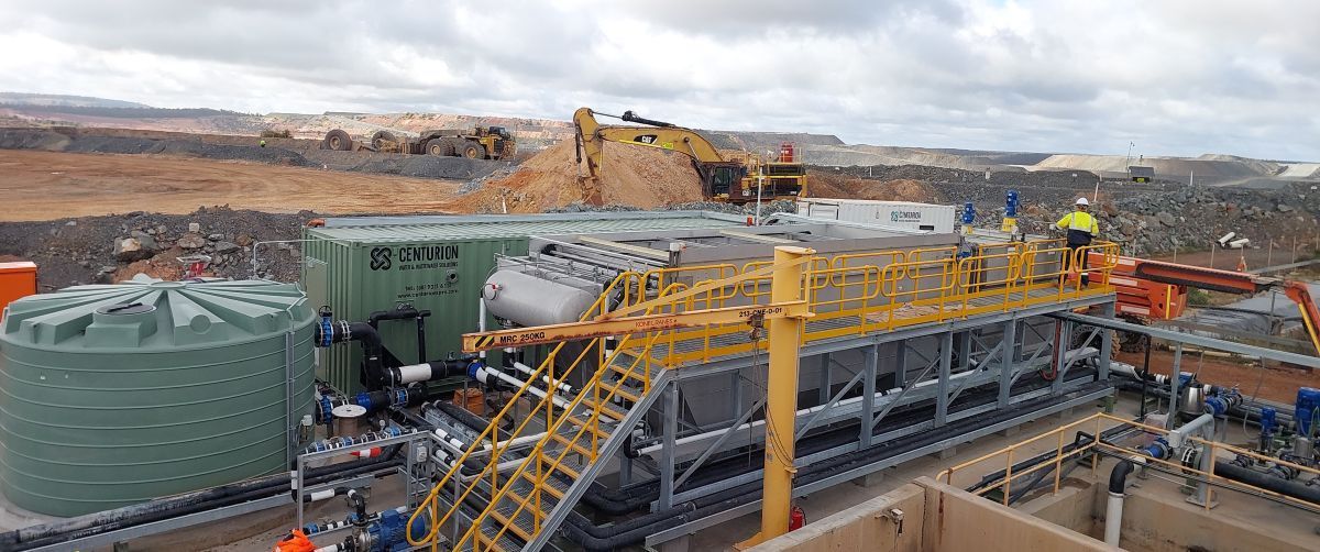 Industrial site with tanks, pipes, and machinery. Workers in high-vis vests and a digger in the background. Overcast sky.