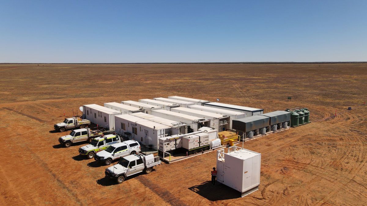 Large white mobile camp with vehicles in a barren red Australian outback under a clear sky.
