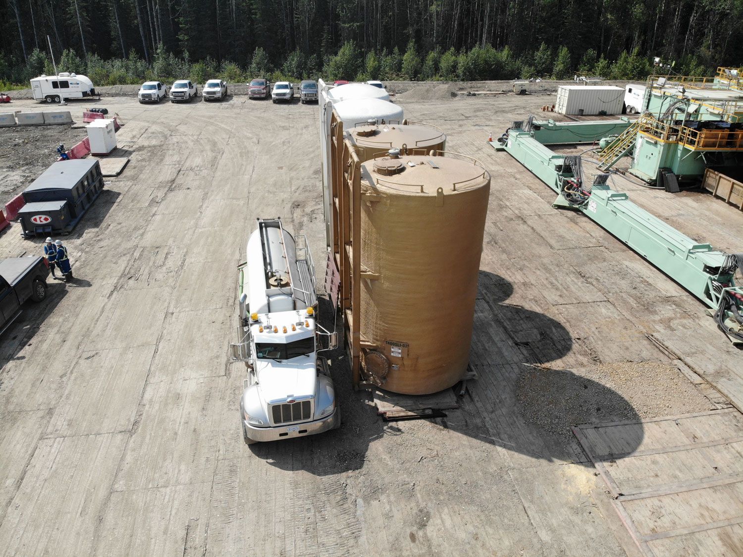 Truck hauling large, fluid management wellsites Canada. Cars and machinery are visible in the background.