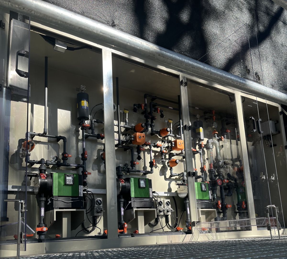 A water treatment system with green pumps, pipes, and control panels inside an open metal enclosure.