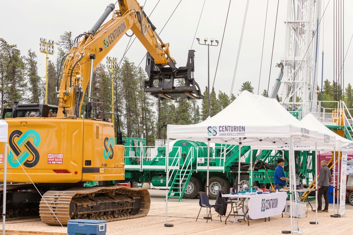 Yellow excavator at an outdoor event beside white tents and green machinery displays