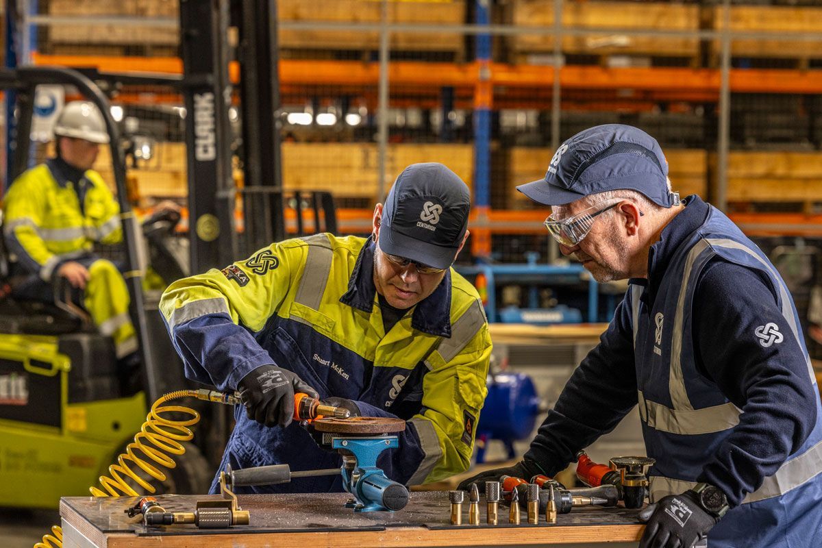 Two workers inspecting and repairing equipment at a warehouse. A forklift operator is in the background.