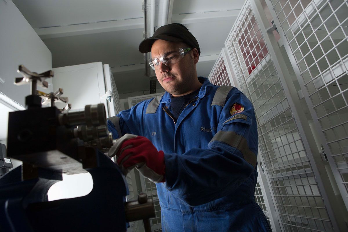 Mechanic in blue coveralls, safety glasses, and gloves working on machinery in a white-walled room.