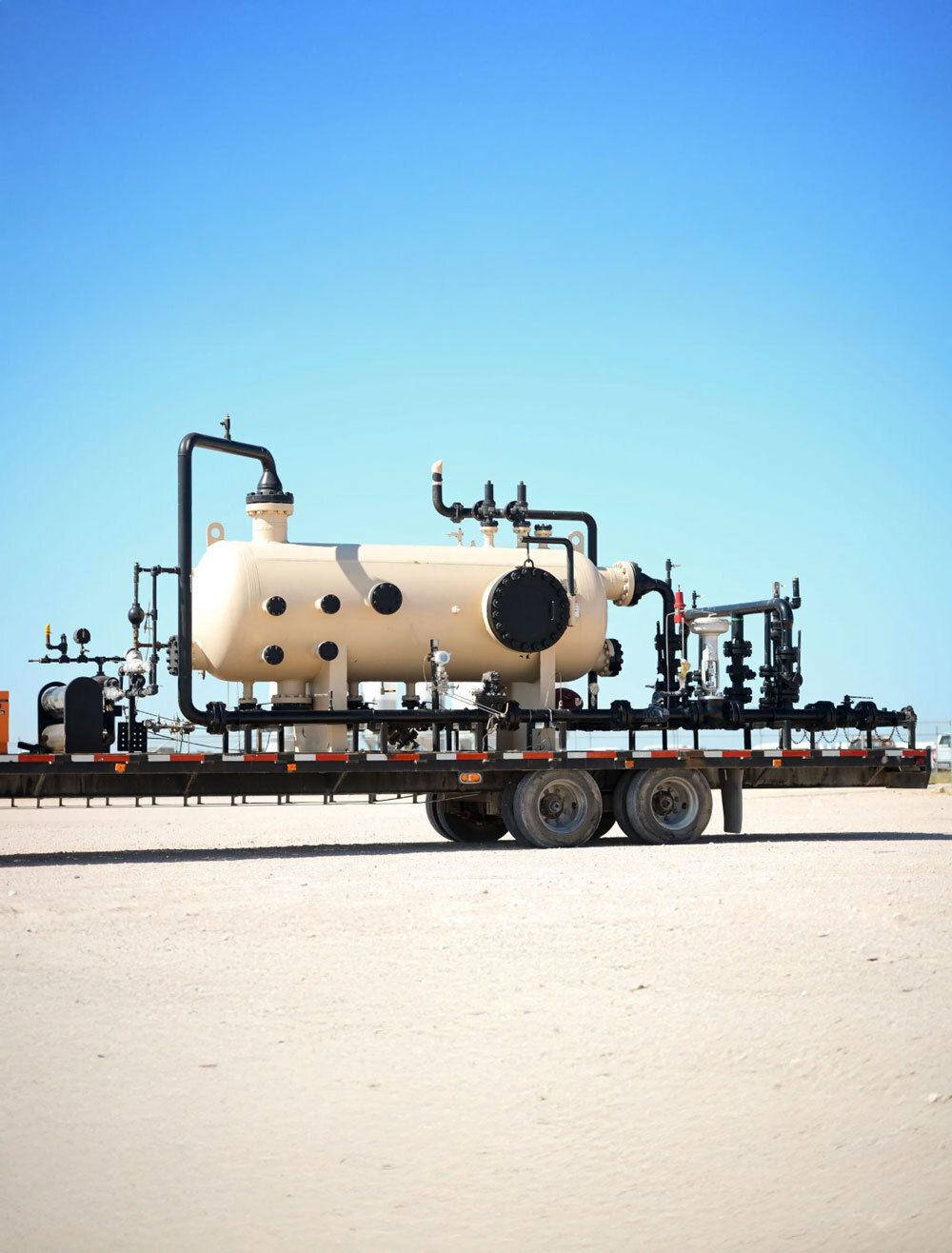 Oilfield separator on trailer, beige tank with black pipes, on a sandy lot against a blue sky.