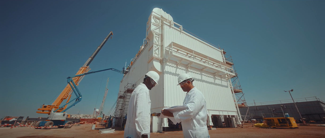 Two workers in white near a large remote instrument enclosure structure under construction.