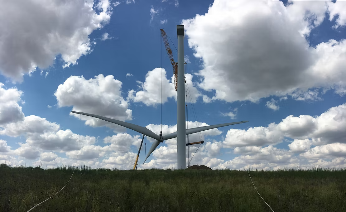 Wind turbine under construction in a grassy field under a cloudy blue sky being hoisted by a crane.