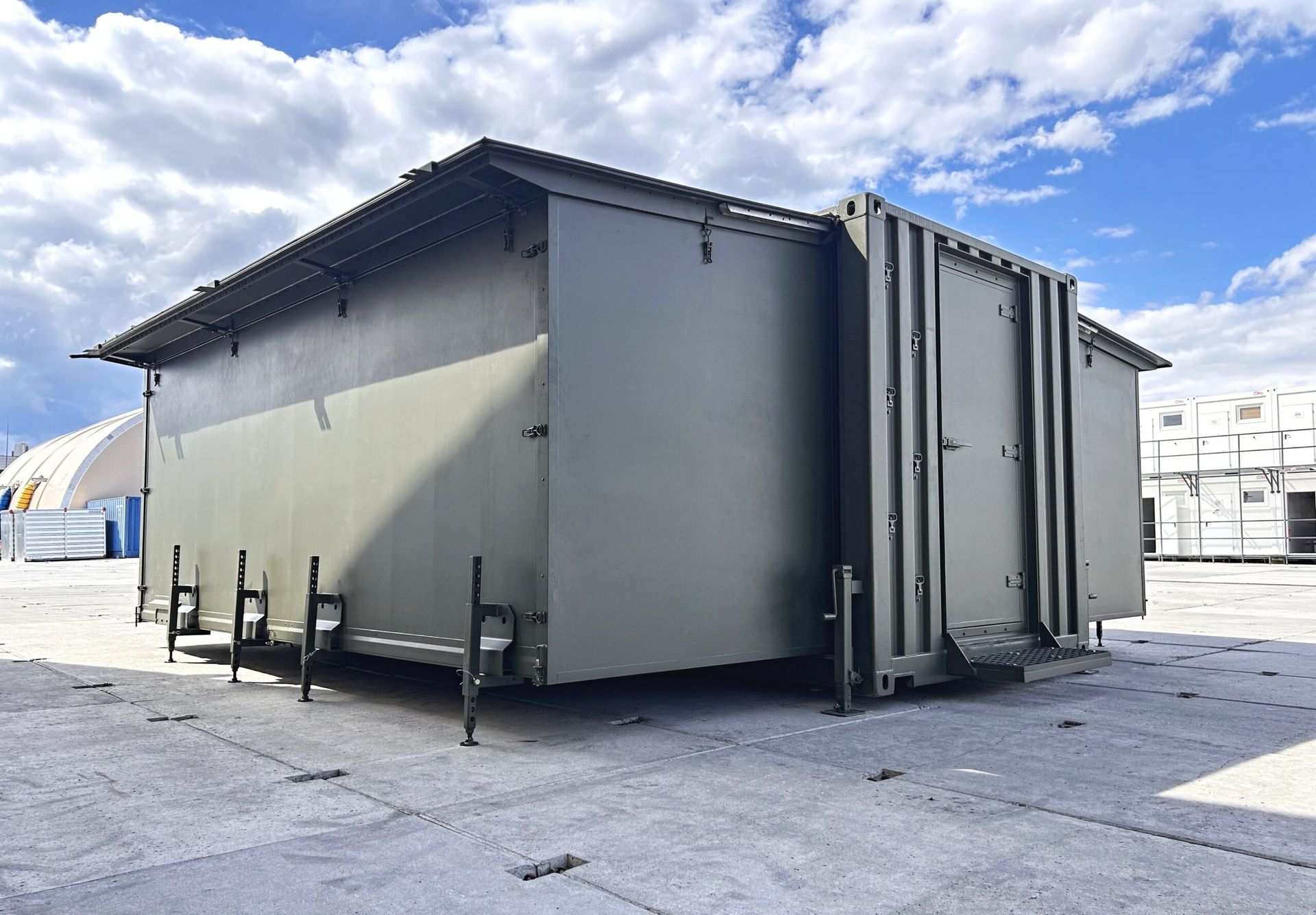 Olive-green, rectangular industrial shelter with roof on a concrete lot, against a blue sky with clouds.