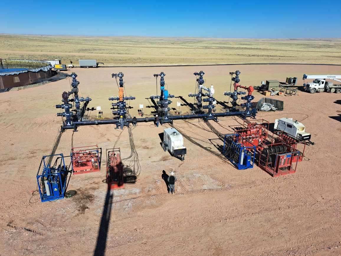 Oil and gas well site with multiple wellheads, pipelines, and equipment, outdoors on a dusty plain under a blue sky.